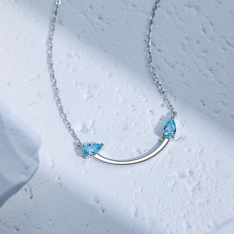 Crafted from polished S925 Sterling Silver, this necklace features a delicate 2.4cm curved bar pendant adorned with shimmering Blue Zircons to mimic a gentle, confident smile. The minimalist design is suspended on an adjustable 40cm clavicle chain, offering a mirror-like finish and a sophisticated touch of quiet luxury.