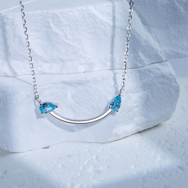 Crafted from polished S925 Sterling Silver, this necklace features a delicate 2.4cm curved bar pendant adorned with shimmering Blue Zircons to mimic a gentle, confident smile. The minimalist design is suspended on an adjustable 40cm clavicle chain, offering a mirror-like finish and a sophisticated touch of quiet luxury.