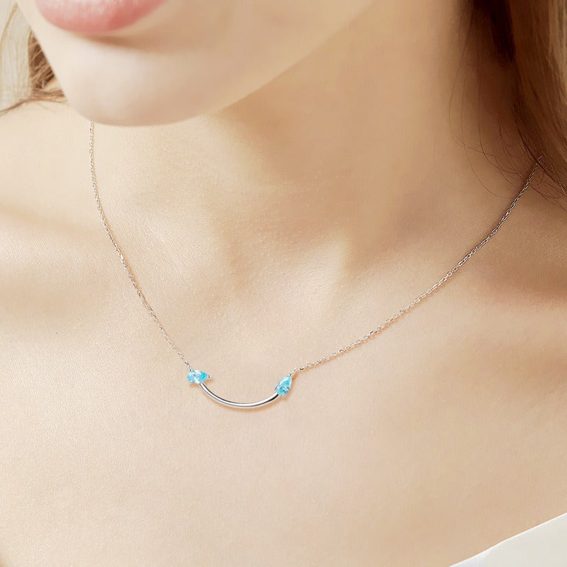 Crafted from polished S925 Sterling Silver, this necklace features a delicate 2.4cm curved bar pendant adorned with shimmering Blue Zircons to mimic a gentle, confident smile. The minimalist design is suspended on an adjustable 40cm clavicle chain, offering a mirror-like finish and a sophisticated touch of quiet luxury.