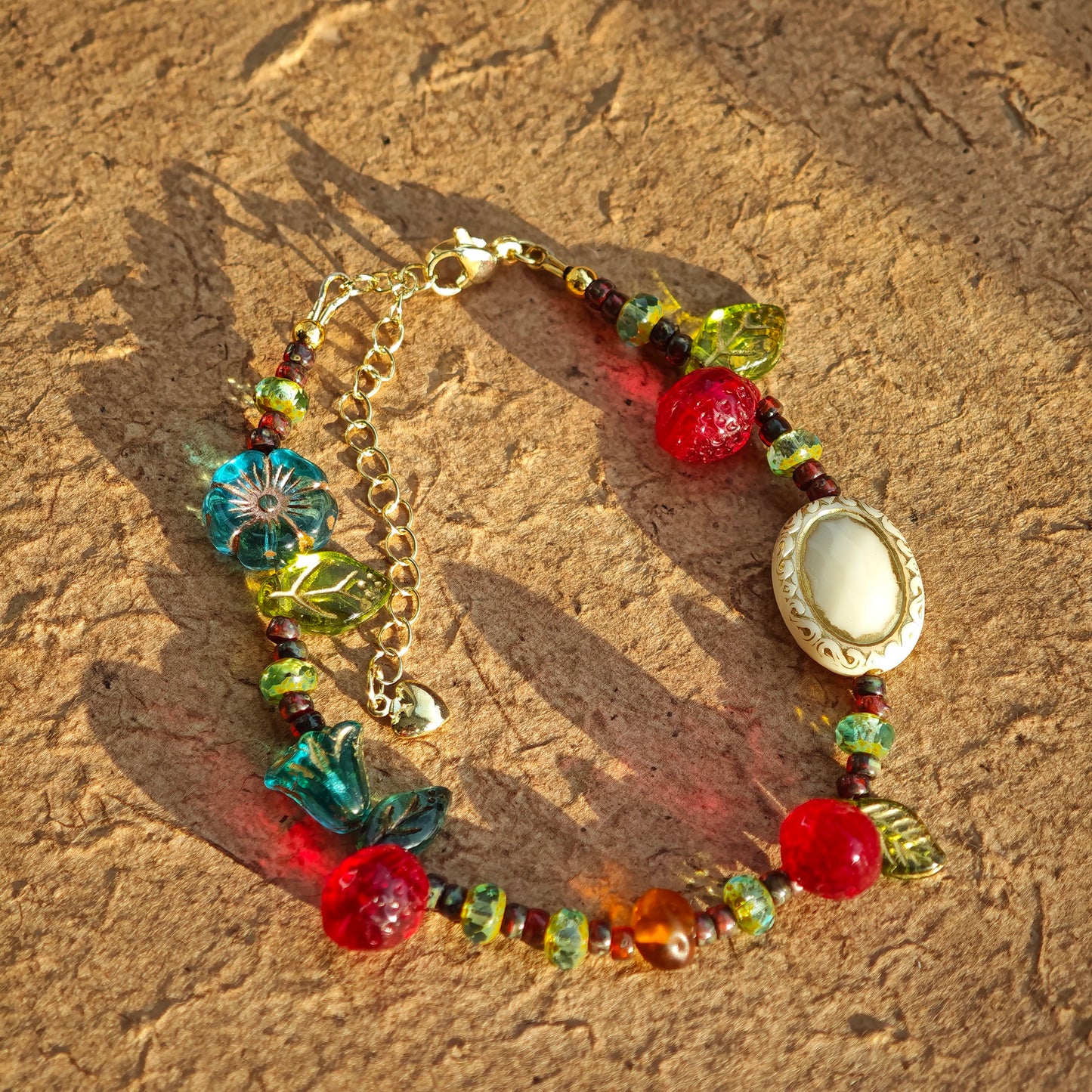 This vibrant bracelet features a colorful mix of ruby-red lemon, sapphire-blue flower, and emerald-green leaf charms, all dangling from a shimmering Czech glass bead chain.