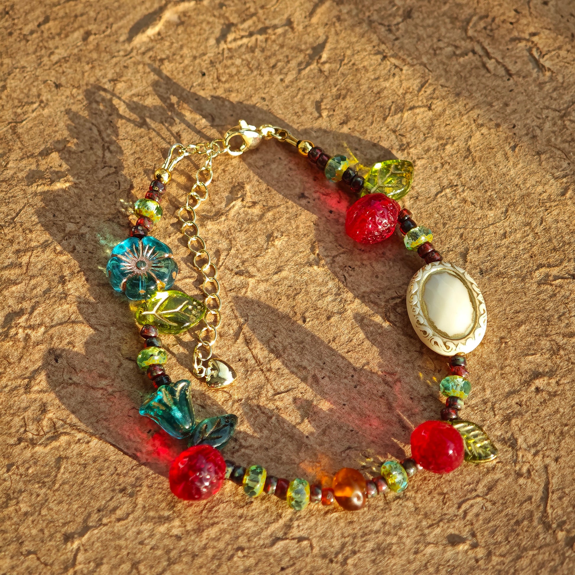 This vibrant bracelet features a colorful mix of ruby-red lemon, sapphire-blue flower, and emerald-green leaf charms, all dangling from a shimmering Czech glass bead chain.