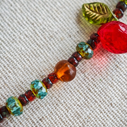 This vibrant bracelet features a colorful mix of ruby-red lemon, sapphire-blue flower, and emerald-green leaf charms, all dangling from a shimmering Czech glass bead chain.