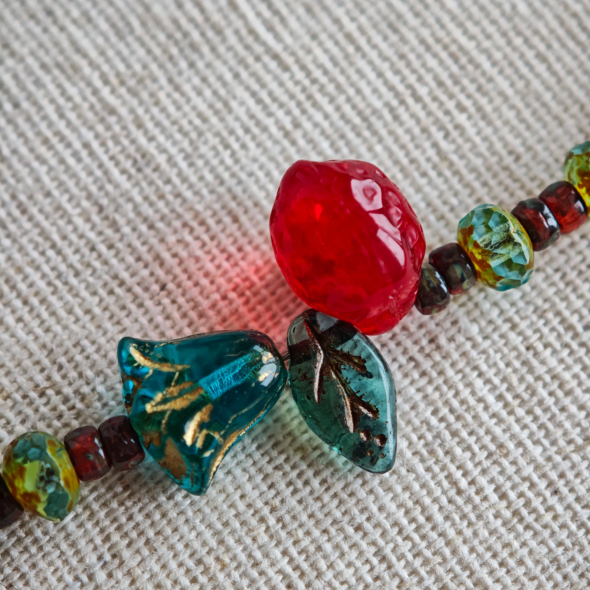 This vibrant bracelet features a colorful mix of ruby-red lemon, sapphire-blue flower, and emerald-green leaf charms, all dangling from a shimmering Czech glass bead chain.
